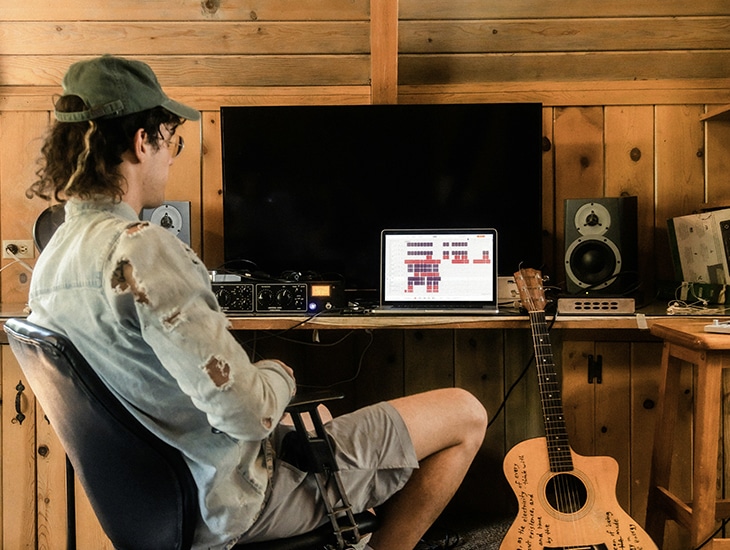 Person in a casual outfit, wearing a hat, sits in front of a computer with audio editing software open. Surrounding the work area are speakers, audio equipment, and an acoustic guitar. A framed Letter from Symphonic's CEO sits proudly on the desk, adding a personal touch to the creative space.